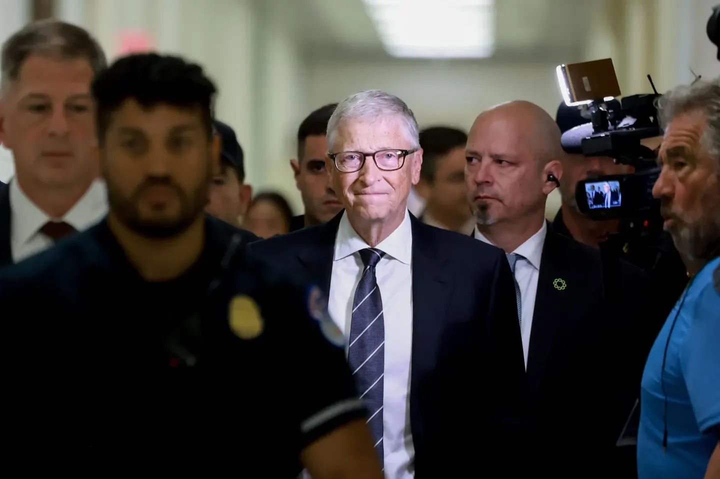 Bill Gates walking through a corridor surrounded by security and cameras
