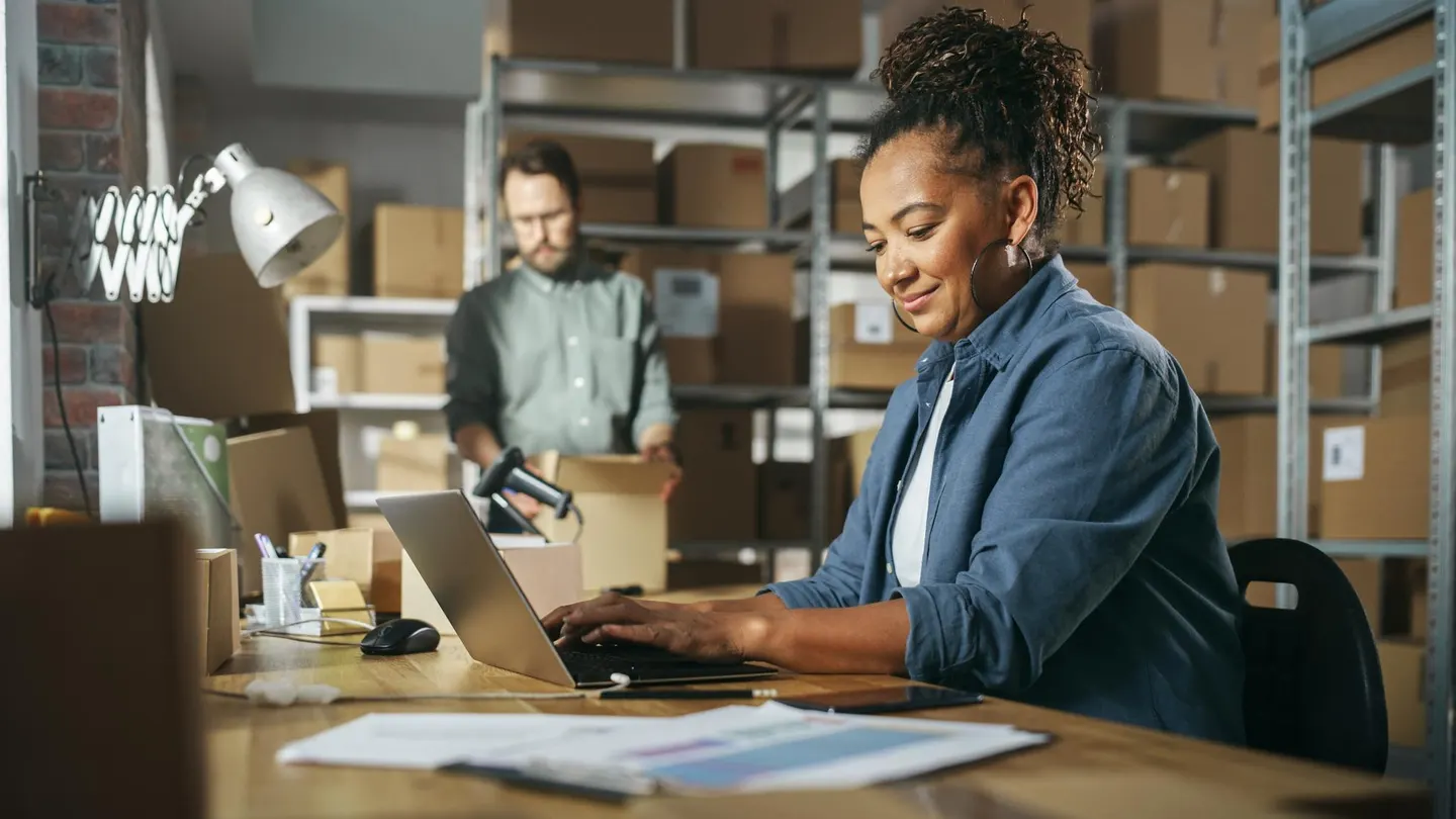 A small business owner working on a laptop in a warehouse while a colleague packs orders in the background