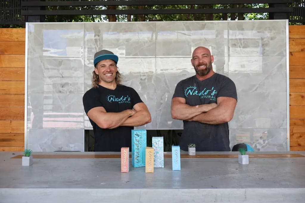 The Mastronardo brothers standing behind a counter lined with Nardo's Natural skincare boxes, arms crossed and smiling